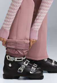 Crne skijaške čizme s više srebrnih kopči, uparene s ružičastim vodootpornim hlačama koje imaju skupljeni elastični rub na gležnju. Bijele rukave s uzorkom vidljive.