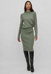Robe pull tricotée verte avec un col montant, texture côtelée et silhouette ajustée, accompagnée de bottes noires brillantes à hauteur de genou.