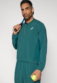 Τζελ αθλητικό μπουφάν με φερμουάρ μπροστά, λεπτομέρειες από δίχτυ και ελαστικές μανσέτες, φορεμένο με ταιριαστό παντελόνι. Κρατώντας ρακέτα τένις και μπάλα.