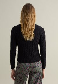 Schwarzer, kabelstrickpullover mit geripptem Saum, langen Ärmeln und taillierter Silhouette, kombiniert mit gemusterten, grünen und rosafarbenen Hosen.