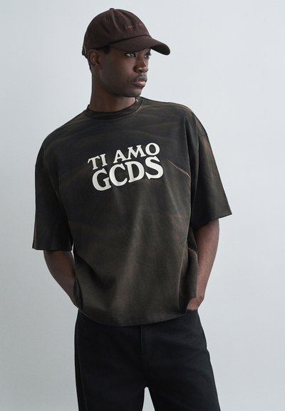T-shirt oversize tie-dye nera e marrone con testo bianco "TI AMO GCDS" al centro, abbinata a pantaloni neri e un cappello marrone.