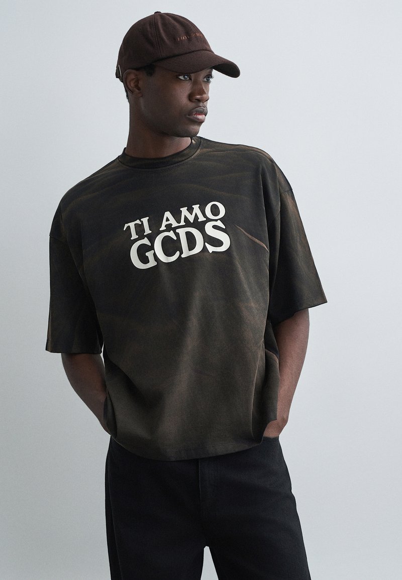 Svart og brun tie-dye oversized t-skjorte med hvit "TI AMO GCDS" tekst i midten, kombinert med svarte bukser og en brun caps.