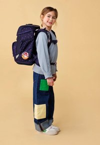 Zaino viola scuro con motivo a stelle e una spilla grafica rotonda; felpa grigia chiara; jeans blu con toppe colorate; sneakers bianche.