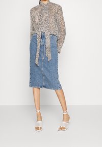 Lys, mønstret bluse i neutrale nuancer med bindebånd, som er parret med en højttaljede denim blyantnederdel og hvide stroppsandaler.