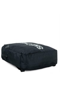 Mochila rectangular negra con una textura suave, que presenta un gran logo blanco en la parte frontal, un bolsillo lateral con cremallera y dos correas ajustables.