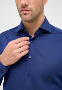 Marineblauwe shirt met een gestructureerd design, contrasterende knopen en een puntige kraag. De drager past met één hand de kraag aan.