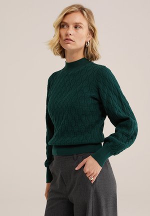 Vrouw met kort blond haar, draagt een donkergroene kabelgebreide trui en grijze broek, kijkt opzij met één hand in de zak.