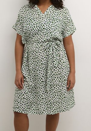 Mujer con un vestido blanco hasta la rodilla con manchas de leopardo verdes y negras y un cinturón de tela a juego atado en la cintura.