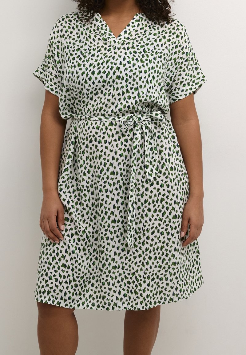 Femme portant une robe blanche mi-longue à taches léopard vertes et noires avec une ceinture en tissu assortie nouée à la taille.