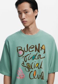 Mint green oversized t-shirt made of soft cotton, featuring artistic brown and colorful text with handprint accents. Short sleeves, crew neck.