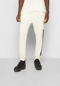 Cream-colored joggers with side cargo pockets, black stripe details, and elastic cuffs, paired with black sneakers.