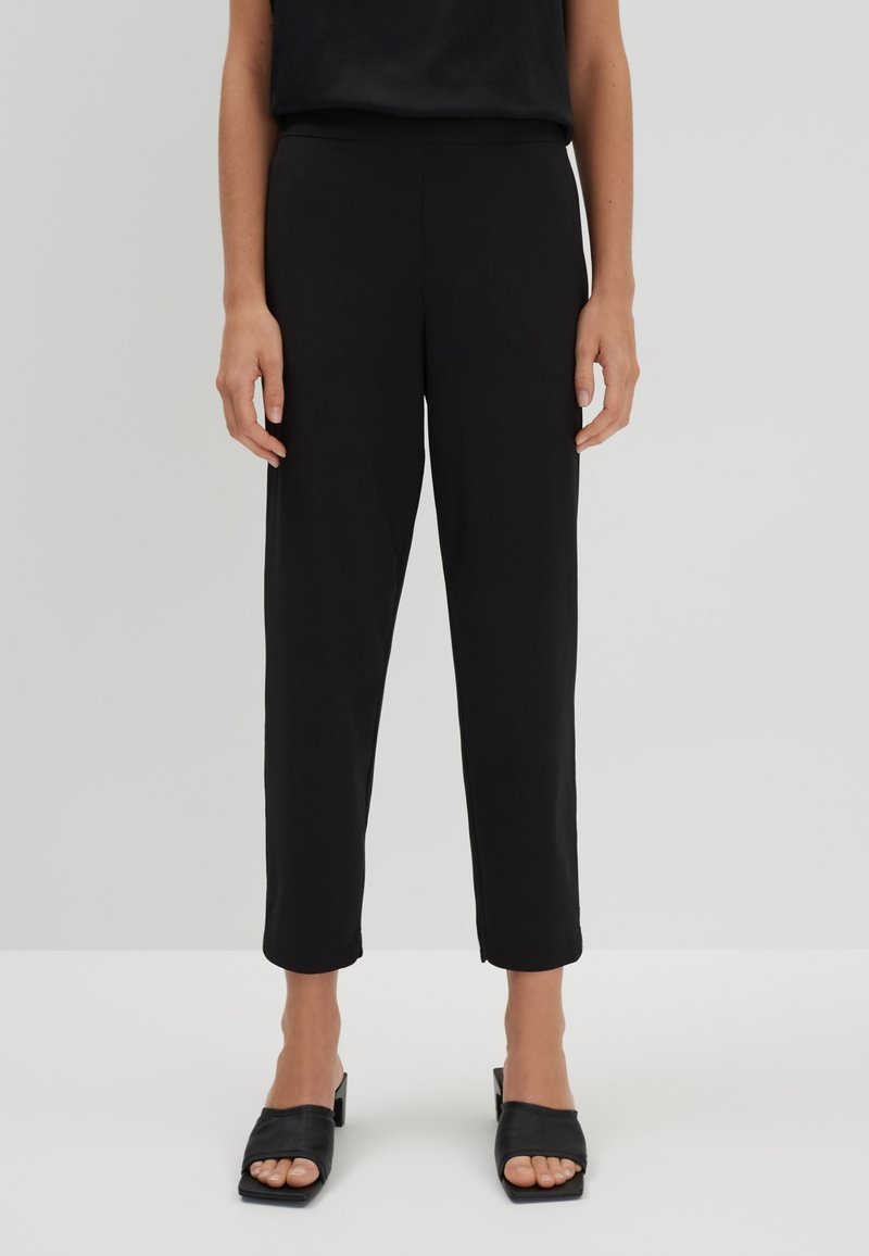 Personne portant un pantalon noir droit court et des sandales noires ouvertes à talons, debout devant un fond uni.