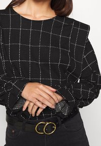 Svart rutig blus med vit rutnät mönster, texturerad tyg, puffärmar och samlade manschetter, tillsammans med ett svart bälte med guldgenomgångar.