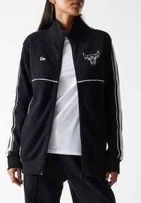 Svart zip-up jakke med hvite kontraststriper, med en okselogo på venstre bryst. Laget av et glatt, strukturert stoff.