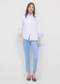 Weißes Hemd mit Knöpfen und gestreiften blauen Manschetten, kombiniert mit hellblauen, maßgeschneiderten Hosen und roten spitzen Absätzen. Glatter Stoff, strukturierte Passform.