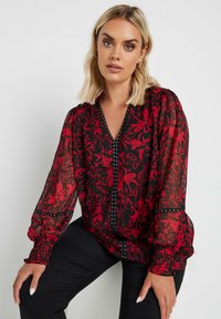 Rote, floral bedruckte transparente Bluse mit V-Ausschnitt, langen Puffärmeln und schwarzen Akzenten. Verfügt über Knopfdetails entlang der Vorderseite.