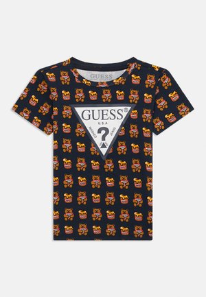 T-shirt en coton bleu marine avec un motif de ours et de pot de miel répété, avec un logo "GUESS" blanc en forme de triangle au centre. Manches courtes.