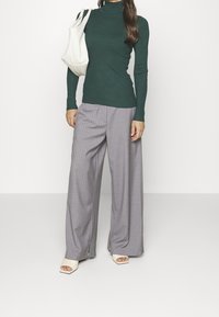 Pull à col roulé côtelé vert, associé à un pantalon large à rayures grises et blanches et des mules à talons ouverts blanches, portant un sac fourre-tout blanc.