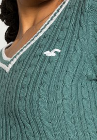 Knitted green sweater with a v-neck, featuring a white striped trim and a small white logo. Textured cable knit pattern throughout.