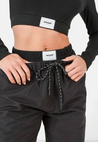 Top negro corto de manga larga con textura acanalada y logo. Pantalones negros con cordón y cinturilla elástica, y cordones blancos en contraste.