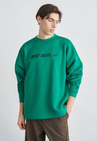 Πράσινο φούτερ με την επιγραφή «JUST DO IT.» σε μωβ γράμματα, στρογγυλή λαιμόκοψη και ριμπ μανσέτες, συνδυασμένο με καφέ παντελόνι.