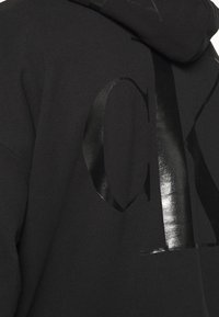 Sudadera negra con capucha que presenta un diseño de logo "CK" negro brillante. La tela es suave, con un aspecto texturizado y un ajuste estándar.