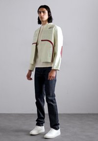 Lyst grøn cropped jakke med præget tekst og røde detaljer, parret med mørke jeans og hvide sneakers på en mandlig model mod en ensfarvet baggrund.