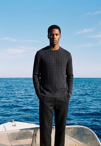 Homme en pull noir et pantalon, debout avec les mains dans les poches sur un bateau, face à un ciel bleu clair et un océan calme en arrière-plan.