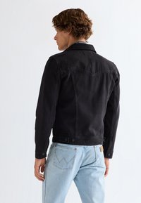 Mand med krøllet brunt hår iført en sort denimjakke og lyseblå jeans, stående med ryggen til mod en ensfarvet hvid baggrund.