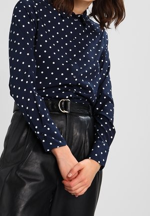 Kobieta ubrana w granatową bluzkę w białe kropki z długim rękawem, wpuszczoną w czarne, skórzane spodnie z wysokim stanem, ozdobione paskiem z okrągłą, srebrną klamrą.