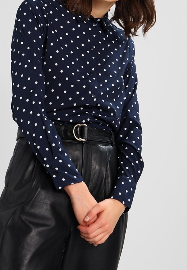 Femme portant un chemisier à manches longues bleu marine à pois, rentré dans un pantalon en cuir noir taille haute avec une ceinture à boucle ronde argentée.