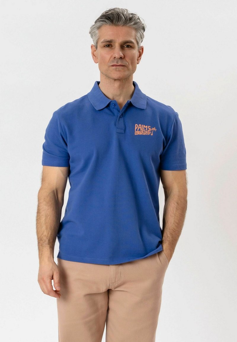 Homme d'âge moyen aux cheveux gris portant un polo bleu avec le logo « Palms Island » et un pantalon beige, debout avec une main dans la poche.