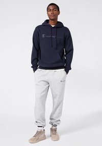 Felpa blu navy con tasca frontale e lacci, abbinata a pantaloni della tuta grigi chiari con logo laterale. Sneakers beige con dettagli testurizzati.