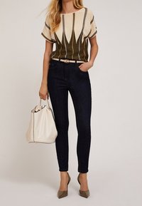 Helle, olivfarbene gemusterte Strickoberteil, kombiniert mit dunklen Jeans. Beiger Gürtel und Handtasche, transparente Absätze. Strukturiertes Material mit grafischen Elementen.