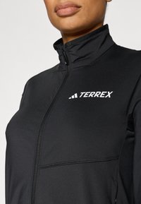 Fekete Terrex dzseki magas gallérral, végig cipzáros elülső résszel és fehér logóval. Sima textúra, testhezálló kialakítás, és látható mintázat nélkül.