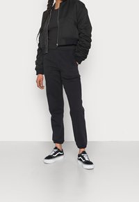 Svart bomberjacka med dragkedja framtill, matchad med avsmalnande svarta byxor och svarta sneakers med vita accenter. Tyget ser mjukt och slätt ut.