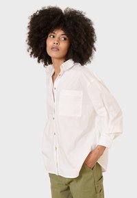 Camicia bianca con bottoni, dal taglio rilassato e una tasca sul petto. Il tessuto appare liscio, con maniche lunghe e colletto a punta.