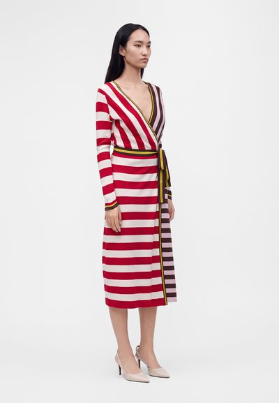 Femme portant une robe à manches longues de style portefeuille avec des rayures horizontales rouges et blanches et des rayures verticales marron contrastantes sur un côté, associée à des talons beiges.