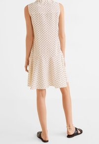 Vestido sin mangas de color beige con lunares negros, con un dobladillo acampanado y una textura suave. Combinado con sandalias planas negras y detalle de tira en el tobillo.