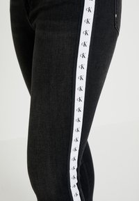 Jeans de mezclilla negros con una raya lateral blanca que presenta el logotipo "cK" repetido. El corte es slim y la textura es suave.
