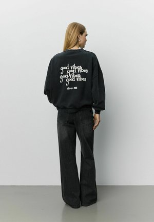 Donna con capelli biondi lunghi che indossa una felpa nera oversize con il testo "good vibes" e jeans neri a gamba larga, in piedi contro un muro semplice.