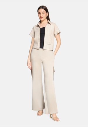 Kvinde iført beige kortærmet jakke med lynlås over sort top, beige bukser med vide ben og sidelommer, samt beige sandaletter med hæl, stående med hånden i lommen.