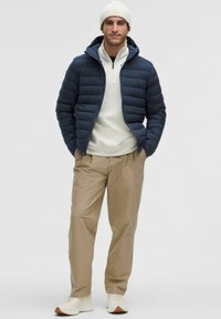 Tengerészkék pufi dzseki vízszintes varrásokkal, fehér, félig cipzáras pulóver, bézs nadrág és világos sportcipők. A viselője kötött sapkát hord.