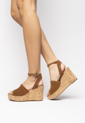 Sandali con zeppa in suede marrone con tacco in sughero. Presentano una cinghia alla caviglia con fibbia e accentuazioni a borchie. Design open toe.