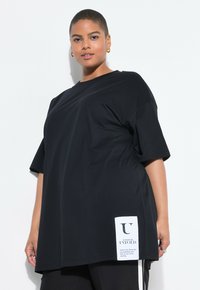 Black oversized t-shirt made from smooth fabric, featuring short sleeves and a side label with text. Cut is loose with a round neckline.