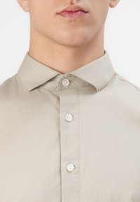 Beige overhemd met knopen, voorzien van een kraag, twee zichtbare knopen bij de hals en een gladde stofstructuur. Minimalistisch ontwerp zonder patronen.