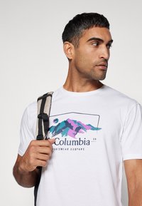 Valkoinen lyhythihainen t-paita, jossa on monivärinen vuoristokuvio ja "Columbia Sportswear Company" -teksti. Yhdistetty harmaaseen reppuun.