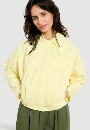 Žena s tmavými vlasmi nosiaca voľnú bledozlutú bundu cez olivovo zelené nohavice, stojaca s rukami vreckách bundy.