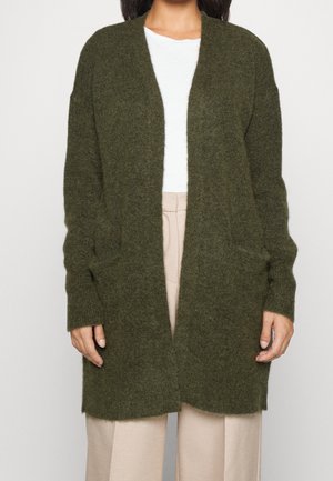 Vrouw draagt een openvallend, lang olijfgroen vest met zakken, gecombineerd met een wit shirt en beige broek.