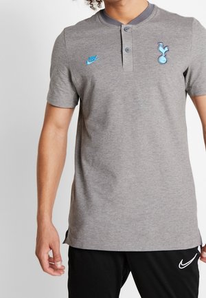 Personne portant un polo gris Nike avec le logo bleu de Tottenham Hotspur et un pantalon noir Nike, debout sur un fond blanc.